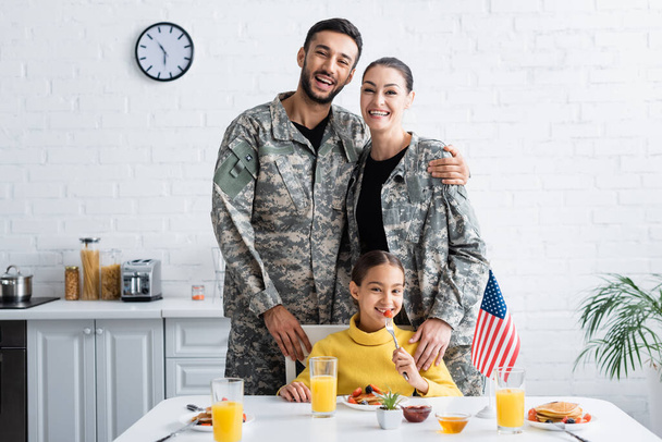 Pais em uniforme militar olhando para a câmera perto da criança, café da manhã e bandeira americana na cozinha  - Foto, Imagem