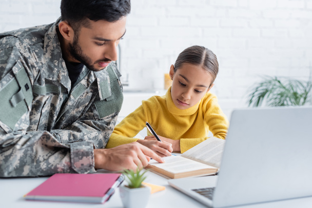 Padre in uniforme militare che punta al libro vicino a figlia e laptop a casa  - Foto, immagini