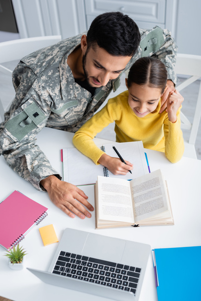 Widok człowieka w mundurze wojskowym siedzącego obok córki piszącego na notatniku i laptopie w domu  - Zdjęcie, obraz