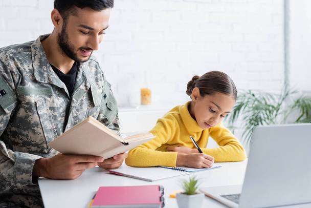 Padre con uniforme militar sosteniendo libro cerca de la hija escribiendo en el cuaderno y portátil en casa  - Foto, imagen