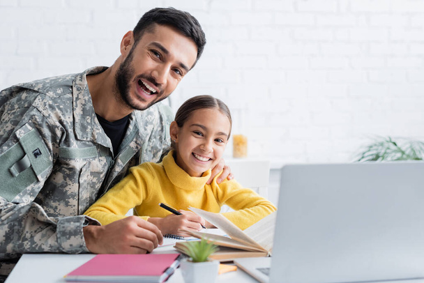 Wesoły mężczyzna w mundurze wojskowym przytulający córkę w pobliżu notebooków i laptopa w domu  - Zdjęcie, obraz
