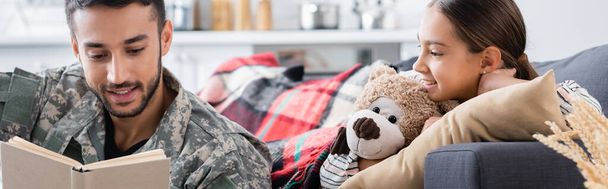Hombre sonriente en uniforme militar libro de lectura cerca de niño con oso de peluche en el sofá, pancarta  - Foto, imagen