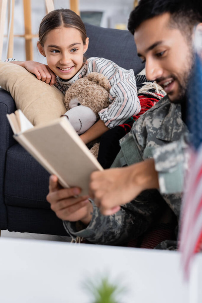 Niño sonriente con oso de peluche mirando al padre borroso en uniforme militar leyendo libro en casa  - Foto, imagen