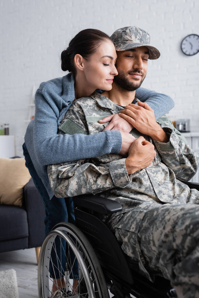 Kamuflaj üniformalı ve tekerlekli sandalyede kocasını kucaklayan bir kadın.  - Fotoğraf, Görsel