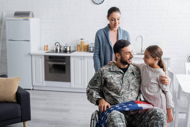 Veterano sonriente en silla de ruedas con bandera americana abrazando a su hija cerca de su esposa en casa  - Foto, imagen