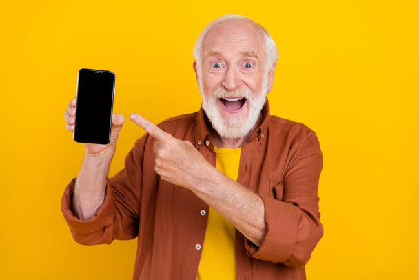 Photo of old man hold phone empty space direct finger touchscreen wear brown shirt isolated yellow color background - 写真・画像