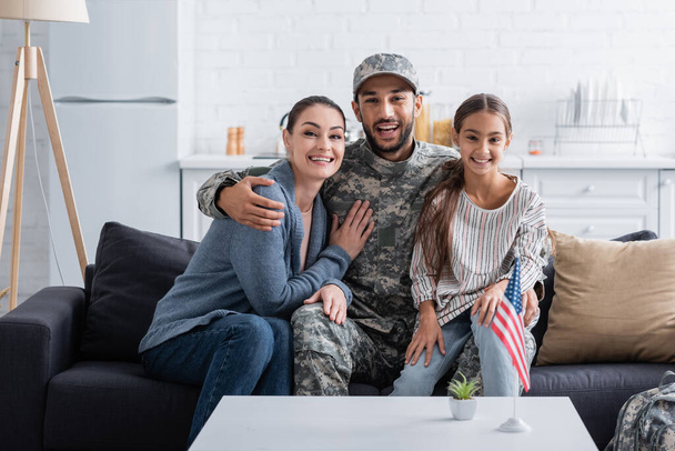 Positive man in camouflage uniform hugging family on couch near american flag at home  - Fotografie, Obrázek