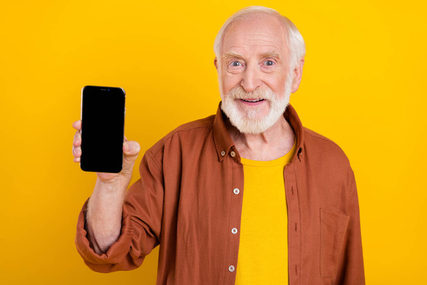 Foto van promotor oude man houd telefoon apparaat tonen lege ruimte scherm slijtage bruin shirt geïsoleerde gele kleur achtergrond - Foto, afbeelding