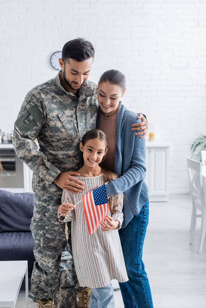 Smiling man in military uniform hugging wife and kid with american flag at home  - Fotoğraf, Görsel
