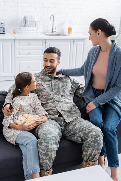 Soldier holding remote controller near daughter with popcorn and wife at home  - Photo, Image