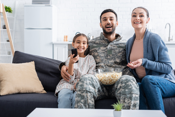 glimlachende familie met popcorn kijken film in de buurt van vader in camouflage uniform thuis  - Foto, afbeelding