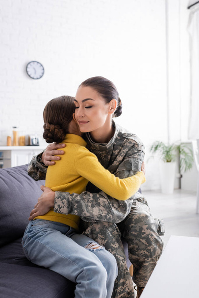 Woman in camouflage uniform hugging daughter on couch at home  - Fotografie, Obrázek