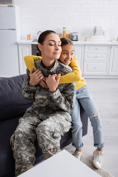 Smiling preteen kid hugging mother in military uniform on couch  - Фото, изображение