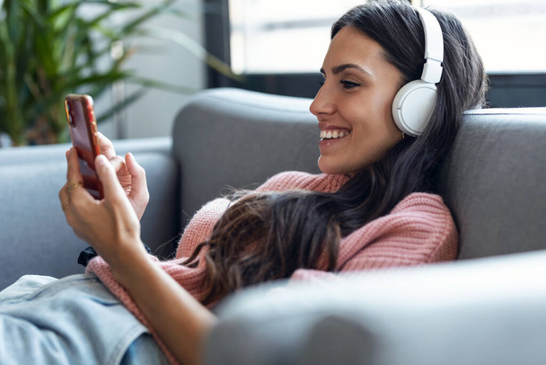Laukaus kaunis nainen kuuntelee musiikkia, kun käytät älypuhelinta istuu sohvalla kotona. - Valokuva, kuva