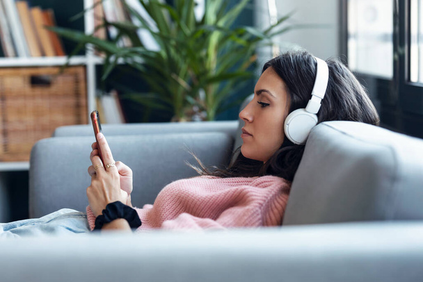 Shot of beautiful woman listening to music while using smartphone sitting on sofa at home. - Foto, immagini