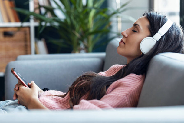 Shot of beautiful woman listening to music while using smartphone sitting on sofa at home. - Fotografie, Obrázek