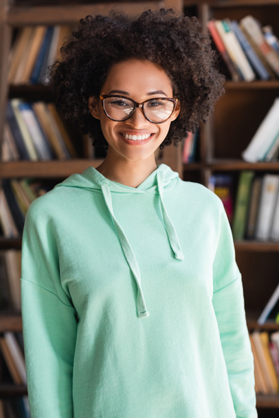 cheerful african american student in eyeglasses looking at camera near bookshelf - 写真・画像