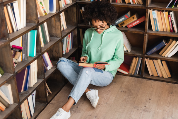 curly african american student in eyeglasses sitting on floor and reading book in library  - 写真・画像