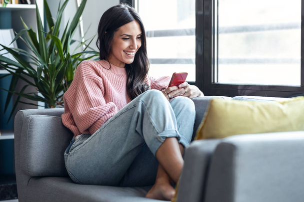 Snímek krásné usměvavé ženy pomocí mobilního telefonu sedí na gauči doma - Fotografie, Obrázek