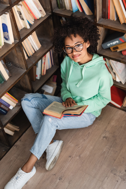 hoge hoek uitzicht van glimlachende Afrikaans-Amerikaanse student in bril zitten met boek in de bibliotheek - Foto, afbeelding