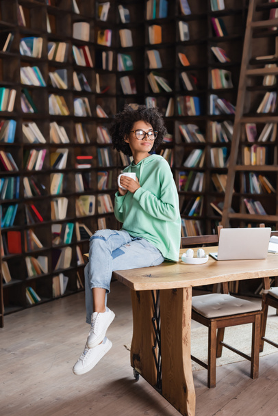 curly african american woman in eyeglasses holding cup of coffee near laptop on desk  - Zdjęcie, obraz