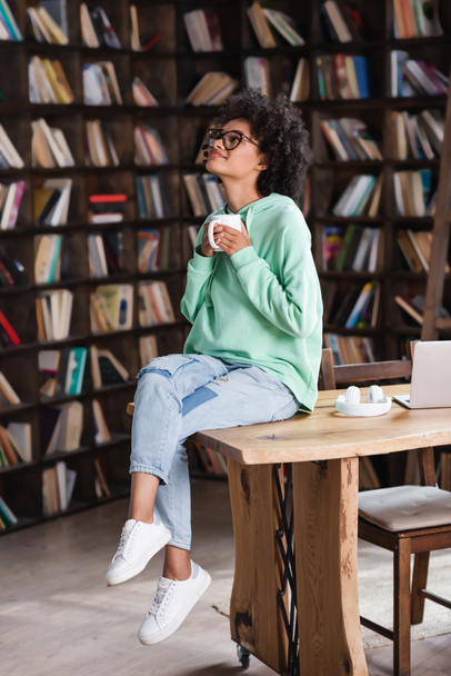 heureux afro-américaine femme dans des lunettes tenant tasse de café près d'un ordinateur portable sur le bureau  - Photo, image