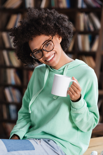 joyful african american woman in eyeglasses holding cup of coffee  - 写真・画像