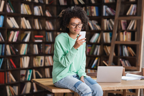 fröhliche afrikanisch-amerikanische Studentin mit Brille, Smartphone in der Hand und am Schreibtisch neben Laptop sitzend  - Foto, Bild