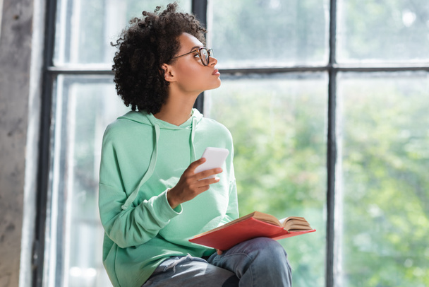 curly african american student in eyeglasses holding cellphone and book while looking at window - Foto, imagen