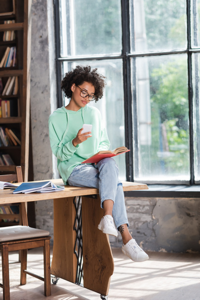 curly african american student in eyeglasses holding cellphone and reading book while sitting on table - Photo, Image