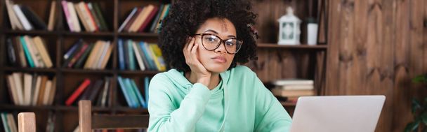 bored african american student in eyeglasses looking at camera while studying online near laptop, banner - Photo, Image