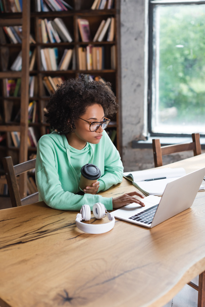 african american student in eyeglasses using laptop while holding paper cup near headphones on desk - 写真・画像