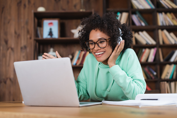 happy african american student in eyeglasses and headphones using laptop - Photo, Image
