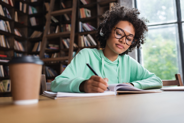 africký americký student v brýlích a sluchátkách psaní v blízkosti notebooku a papírového kelímku - Fotografie, Obrázek