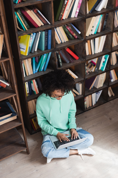 high angle view of curly african american student in eyeglasses using laptop while sitting on floor in library - Foto, Imagen