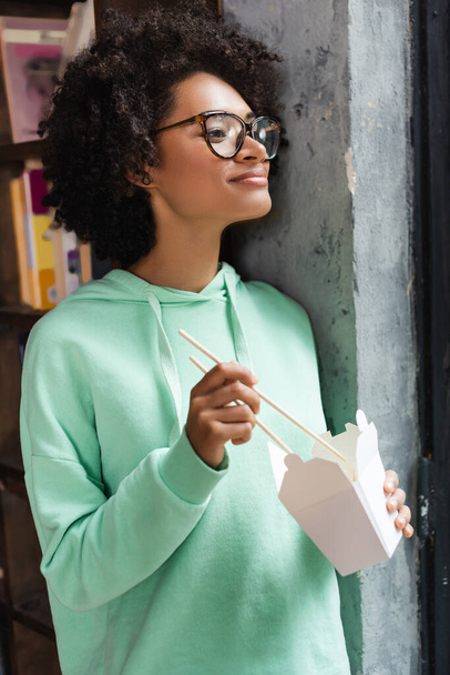 mulher americana africana satisfeito em óculos segurando pauzinhos perto caixa de papelão com takeaway almoço  - Foto, Imagem