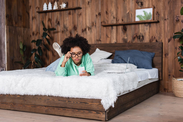 africká americká žena nastavuje brýle a leží na posteli při použití smartphonu v blízkosti notebooku  - Fotografie, Obrázek