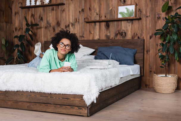 Femme afro-américaine coûteuse dans des lunettes allongées sur le lit tout en utilisant un smartphone près d'un ordinateur portable - Photo, image
