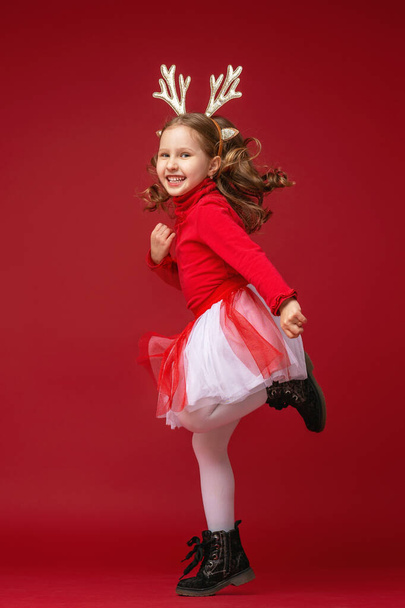 happy funny little girl in Golden reindeer antlers running and jumping on red background in Studio on Christmas day. The child runs and jumps in full growth. Dynamic image. Copy space. - Fotografie, Obrázek