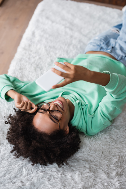 Hochwinkelaufnahme einer glücklichen Afroamerikanerin mit Brille, die auf dem Bett liegt und ein Selfie macht  - Foto, Bild