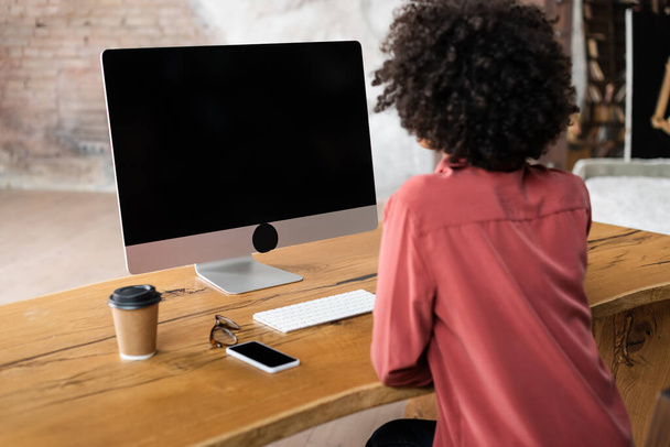 vissza kilátás göndör afro-amerikai nő számítógép közelében okostelefon, papír pohár és szemüveg az asztalon - Fotó, kép