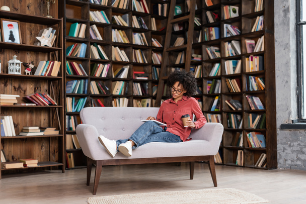 curly african american woman holding paper cup while using digital tablet near bookshelf  - Foto, afbeelding