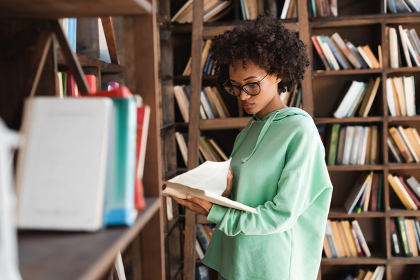 jovem estudante afro-americano em óculos lendo livro perto de estante borrada em primeiro plano - Foto, Imagem