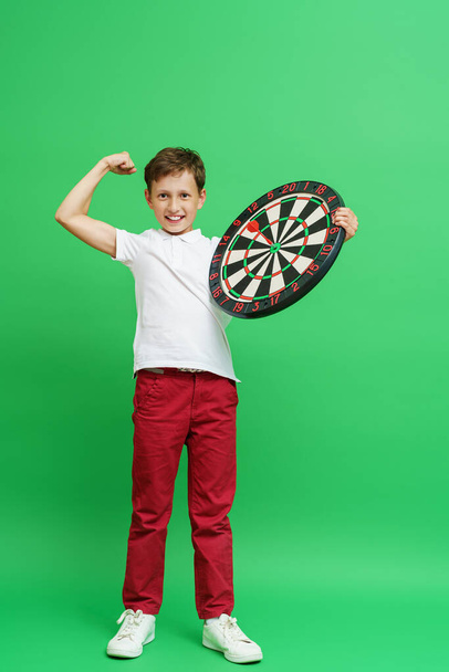 Happy emotional boy of 8 years old, smiling and holding a dart board with a dart in the center standing full-length on a green background. child rejoices in the victory in darts, hit the bull's-eye. - Foto, immagini