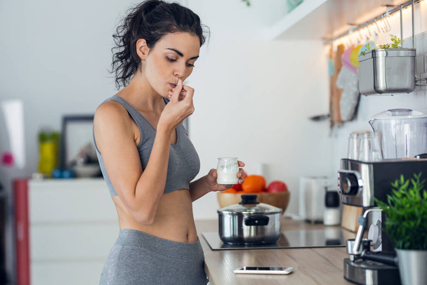 Tiro de mujer deportiva joven comiendo yogur mientras se utiliza el teléfono móvil de pie en la cocina en casa. - Foto, Imagen