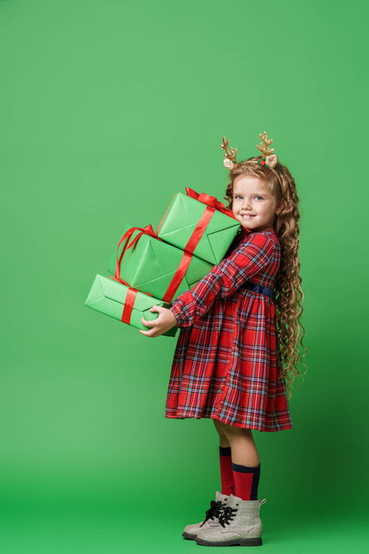 cute little girl of 7 years old with curly hair in golden deer horns, sitting with gift boxes on a green background in the studio. the child smiles happily and looks into the frame. Advertising. - Foto, afbeelding