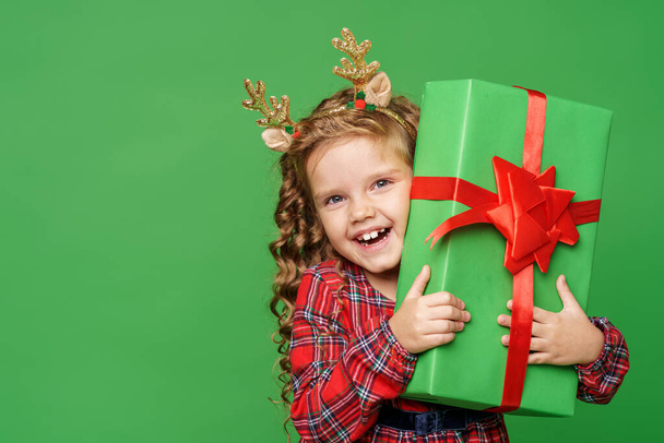 cute little girl of 7 years old with curly hair in golden deer horns, standing with gift boxes on a green background in studio. child smiles happily and looks into the frame. Advertising. copy space - Fotoğraf, Görsel