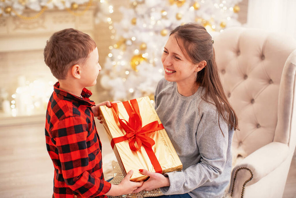 Merry Christmas and Happy Holidays. Happy mom and son hug and hold a Christmas gift box on the background of a Christmas tree decorated with a garland. Copy space - Фото, зображення