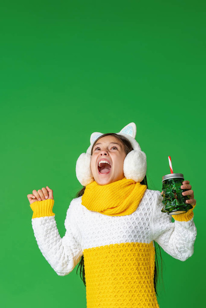 happy teenage girl in knitted sweater, fur headphones on her head with a cocktail glass with a straw, walks on green background. Preparing for Christmas - Photo, Image