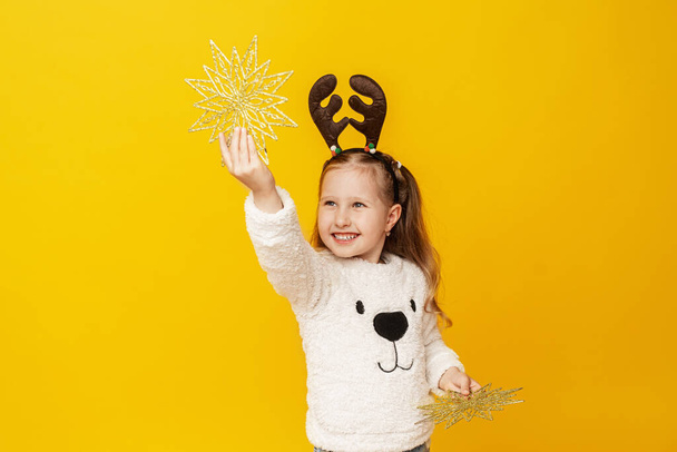 Happy little girl with deer antlers with Christmas stars on yellow background. child is happy, having fun and preparing for Christmas. Advertising. Copy space. - Valokuva, kuva